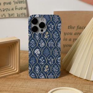 May include: A navy blue phone case featuring a floral design in white, light blue, and yellow. The case has a gold-coloured arched pattern. The phone case has a camera cutout and is on a wooden surface.