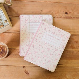May include: Two composition books featuring a light pink and white pattern of small bows and floral sprigs. Each book has a central white rectangular label with the text COMPOSITION BOOK printed in a simple font on the front cover.