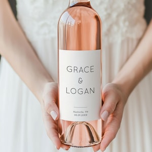 Puede incluir: Una botella de vino rosado con una etiqueta blanca con los nombres "GRACE & LOGAN" y la fecha "08.09.20XX" con la ubicación "Nashville, TN". La botella es sostenida por una persona que lleva un vestido blanco.