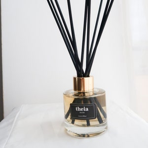 Op de afbeelding: Een heldere glazen fles met een gouden dop en zwarte stokjes. De fles bevat een lichtbruine vloeistof en heeft een zwart etiket met de tekst "theia orchid luxury diffuser".