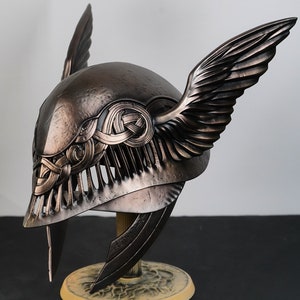 May include: A bronze helmet with wings and intricate detailing. The helmet is on a decorative stand.