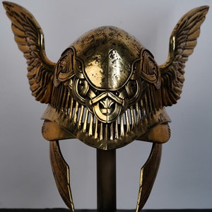 May include: A gold helmet with wings and intricate detailing. The helmet is on a stand and has a textured surface.