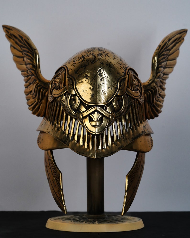 May include: A gold helmet with wings and intricate detailing. The helmet is on a stand and has a textured surface.