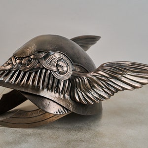May include: A bronze helmet with a winged design. The helmet has a detailed, intricate pattern on the front and is decorated with a Celtic knot design.