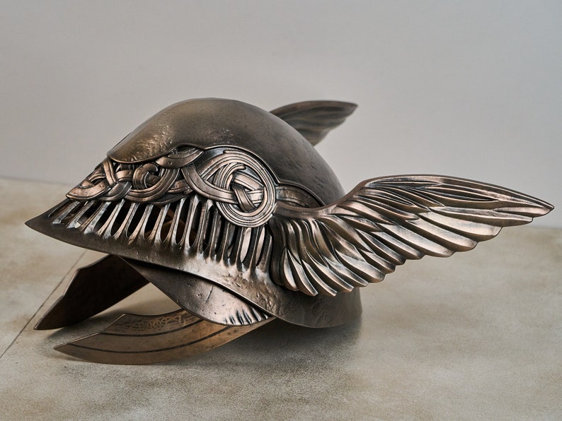 May include: A bronze helmet with a winged design. The helmet has a detailed, intricate pattern on the front and is decorated with a Celtic knot design.