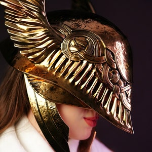 May include: A gold helmet with intricate details and a winged design. The helmet is made of metal and has a textured surface.