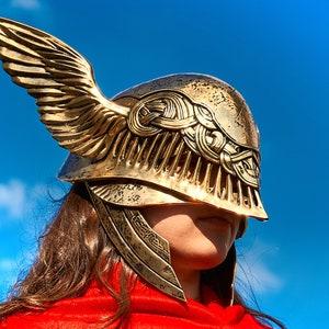 May include: A gold helmet with a winged design. The helmet has intricate details and a decorative pattern. The helmet is worn by a person with long brown hair.