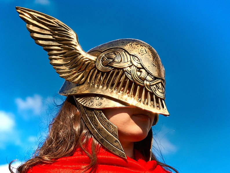 May include: A gold helmet with a winged design. The helmet has intricate details and a decorative pattern. The helmet is worn by a person with long brown hair.