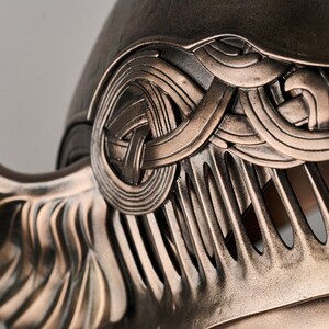 May include: A bronze helmet with intricate details and a winged design. The helmet has a textured surface and a decorative pattern of interwoven lines.