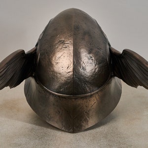 May include: A bronze helmet with wings on the sides. The helmet is smooth and has a textured surface.