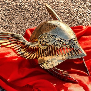 May include: A gold helmet with wings, decorated with intricate designs, resting on a red fabric background.