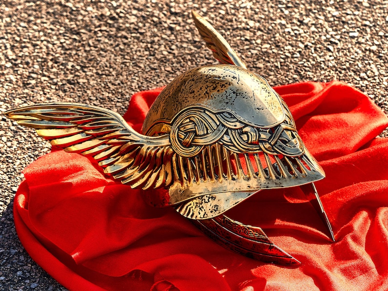 May include: A gold helmet with wings, decorated with intricate designs, resting on a red fabric background.