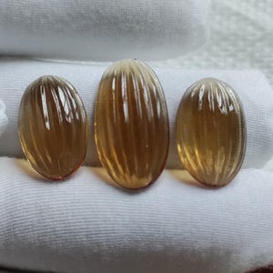 May include: Three oval-shaped, amber-colored gemstones with vertical ridged detailing. The gemstones are displayed on a white surface, showcasing their translucent quality and warm tones. The stones vary slightly in size.