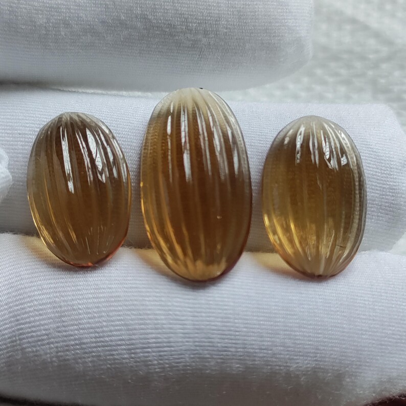 May include: Three oval-shaped, amber-colored gemstones with vertical ridged detailing. The gemstones are displayed on a white surface, showcasing their translucent quality and warm tones. The stones vary slightly in size.