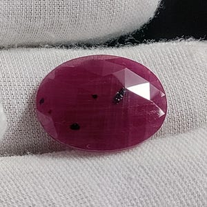 May include: An oval-shaped, faceted ruby gemstone with a deep magenta-red colour. The surface of the stone features several small, distinct black inclusions scattered across its top, and the cut creates a series of geometric, light-reflecting facets.