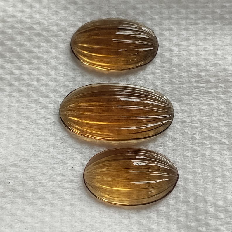 May include: Three oval-shaped, amber-colored gemstones with a ribbed texture. The gemstones are arranged vertically against a white background. The gems have a polished, translucent appearance, reflecting light.
