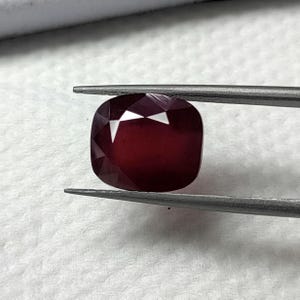 May include: A deep red gemstone with a cushion cut, held by tweezers.