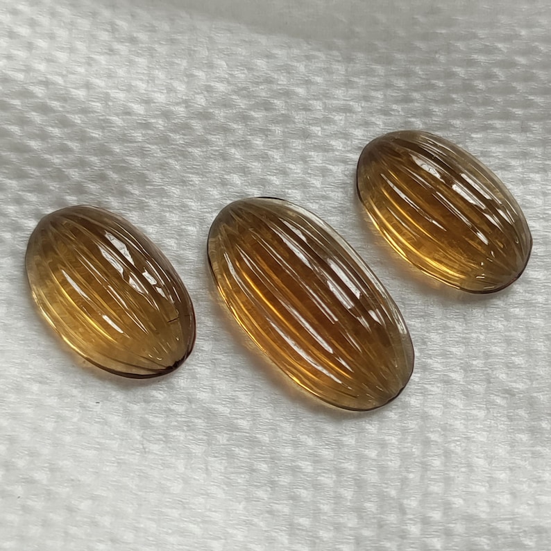 May include: Three oval-shaped, amber-colored gemstones with a ribbed surface. The gems are arranged on a white, textured surface. The stones have a polished, translucent appearance, reflecting light.