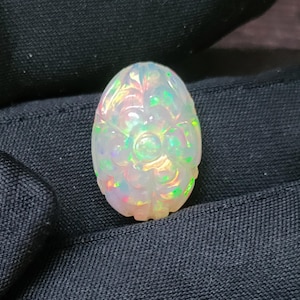 May include: An oval-shaped, carved opal gemstone. The opal is white with iridescent flashes of green, pink, and yellow. The carving depicts a floral design with a central flower and surrounding leaves. The gemstone is set against a dark background.
