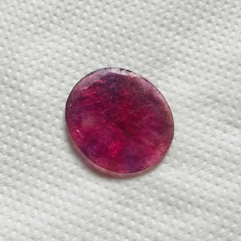 Puede incluir: Una piedra preciosa redonda, facetada, p&uacute;rpura y rosa con una superficie lisa y pulida.