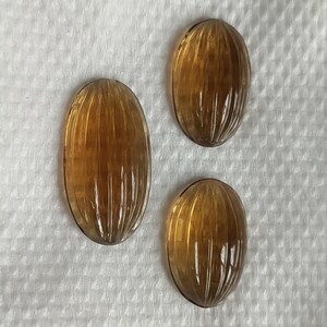 May include: Three oval-shaped, amber-colored gemstones with carved vertical lines. The gemstones are arranged on a white, textured surface. The carvings create a ribbed effect, enhancing the light refraction.