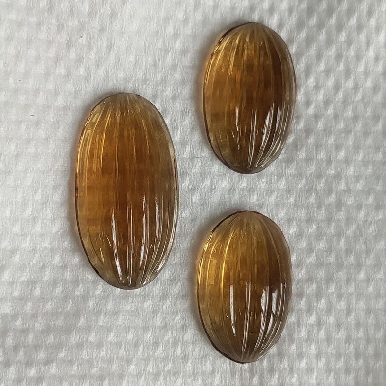 May include: Three oval-shaped, amber-colored gemstones with carved vertical lines. The gemstones are arranged on a white, textured surface. The carvings create a ribbed effect, enhancing the light refraction.