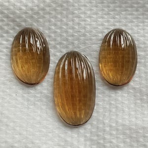 May include: Three oval-shaped gemstones with a warm amber hue. Each gem features a series of vertical, parallel grooves, creating a textured surface. The gems are displayed on a white, textured background.