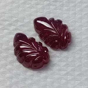 Op de afbeelding: Twee gepolijste, bladvormige edelstenen in een diepe bordeauxrode kleur. De edelstenen hebben gedetailleerde, gesneden aderen en een glanzende afwerking. De achtergrond is een getextureerd, gebroken witte oppervlak.
