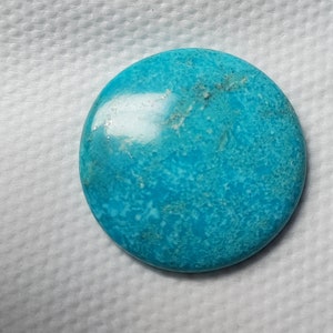 May include: A circular turquoise gemstone, polished to a smooth finish. The stone displays a bright blue colour with natural variations in texture and tone. This gemstone is likely intended for use in jewellery or as a decorative piece.