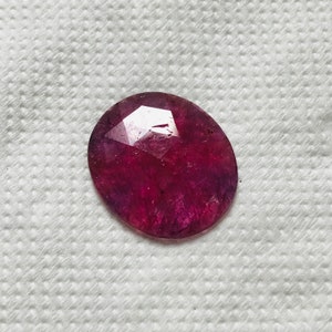 Puede incluir: Una piedra preciosa ovalada de color rojo oscuro con un corte facetado. La piedra preciosa est&aacute; colocada sobre un fondo blanco.