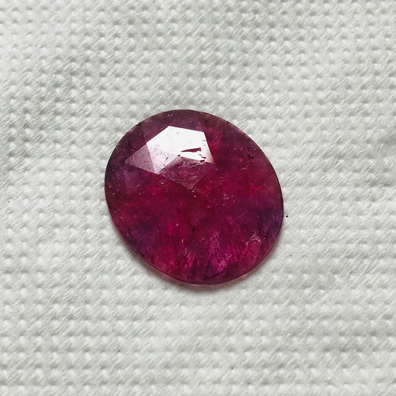 Puede incluir: Una piedra preciosa ovalada de color rojo oscuro con un corte facetado. La piedra preciosa est&aacute; colocada sobre un fondo blanco.