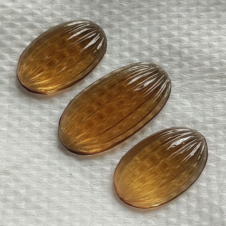 May include: Three amber-colored, oval-shaped gemstones with a ribbed texture and a polished finish. The gemstones are arranged on a white, textured surface, showcasing their translucent quality and intricate design.