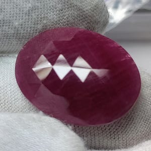 Pode incluir: Uma pedra preciosa de formato oval, na cor magenta profunda, com uma superfície facetada. A pedra exibe um padrão distinto de três reflexos de luz triangulares brilhantes no centro, destacando o corte polido e multifacetado da gema.