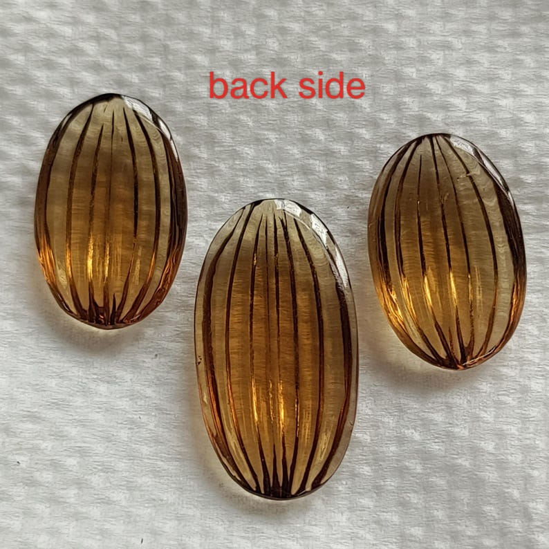 May include: Three oval-shaped, amber-colored glass beads with vertical ribbed detailing. The beads are arranged on a white surface, with the text "back side" in red at the top. The beads are translucent, allowing light to pass through.