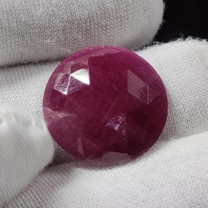 May include: A round, faceted gemstone with a deep magenta-red color. The stone features a polished surface with multiple geometric facets that reflect light, creating a sparkling appearance across its circular, translucent body.
