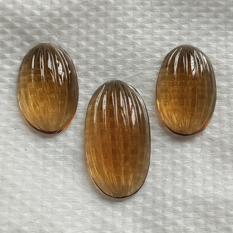 May include: Three oval-shaped gemstones with a warm amber hue. Each gem features a series of vertical, parallel grooves, creating a textured surface. The gems are displayed on a white, textured background.