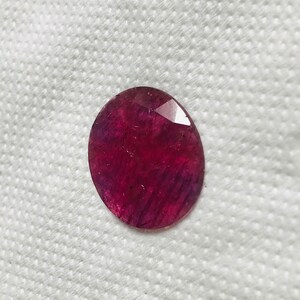 Puede incluir: Una piedra preciosa ovalada de color rojo intenso con un corte facetado. La piedra preciosa tiene un ligero tono p&uacute;rpura y est&aacute; colocada sobre un fondo blanco.
