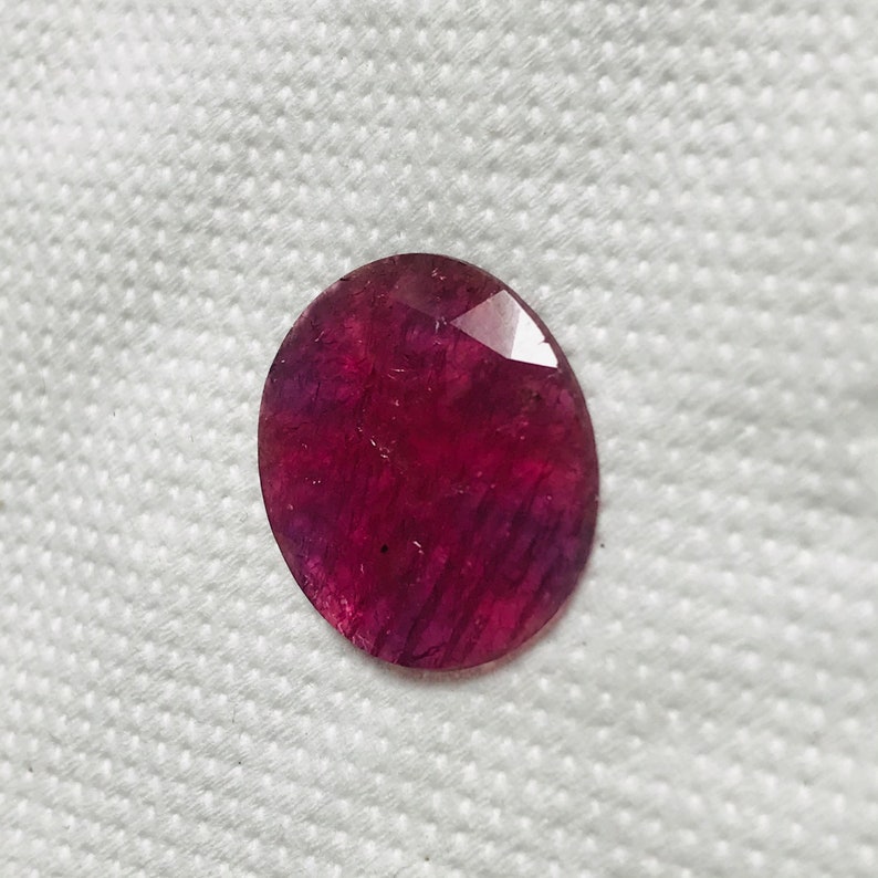 Puede incluir: Una piedra preciosa ovalada de color rojo intenso con un corte facetado. La piedra preciosa tiene un ligero tono p&uacute;rpura y est&aacute; colocada sobre un fondo blanco.