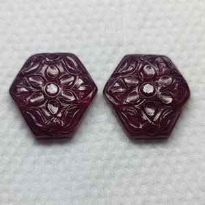 Op de afbeelding: Twee gesneden robijn edelstenen in een zeshoekige vorm met een bloemmotief. De edelstenen zijn diep rood.