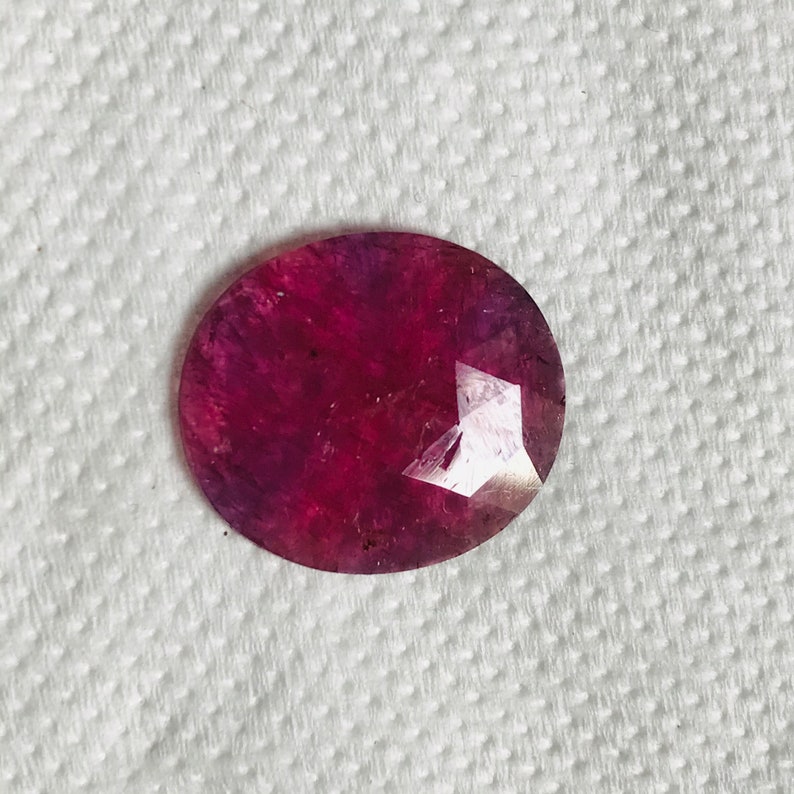 Puede incluir: Una piedra preciosa ovalada de color rojo intenso con un corte facetado. La piedra preciosa tiene un ligero tono p&uacute;rpura y se coloca sobre un fondo blanco.