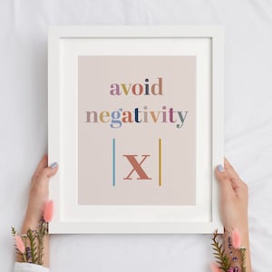 May include: White framed art print with the words "avoid negativity" in colorful text. The print also features the letter "X" with vertical lines on either side. The art is held by hands.