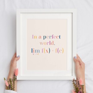May include: White framed art print with the text "In a perfect world, lim f(x) = f(c)" in colorful letters. The print is held by hands with floral accents, set against a white background.
