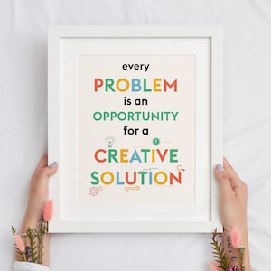 May include: White framed art print with the text "every PROBLEM is an OPPORTUNITY for a CREATIVE SOLUTION" in colorful block letters. The print is held by hands, with floral accents.