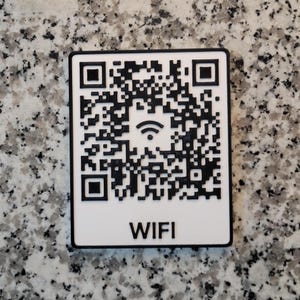May include: A rectangular white sign with a black border featuring a black QR code pattern. In the center of the QR code is a black Wi-Fi signal icon. Below the QR code, the word WIFI is printed in bold, black, sans-serif capital letters.