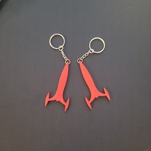 May include: Two red keychains with a stylized, wing-like design. Each keychain has a silver-colored ring and chain. The keychains are displayed on a dark gray surface.