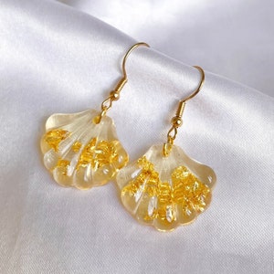 May include: Gold shell earrings with gold flakes embedded in the clear resin. The earrings are on a white background.