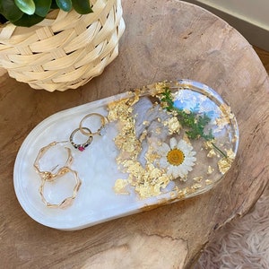 Könnte beinhalten: Ein weißes, ovales Tablett aus Harz mit goldenen Blatt-Akzenten und einer gepressten weißen Blume. Das Tablett ist mit Blattgold und einer gepressten weißen Blume gefüllt. Auf der linken Seite des Tabletts befinden sich drei goldene Ringe.