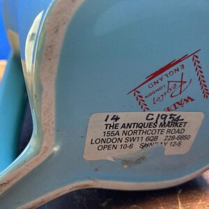 May include: A light blue ceramic pitcher with a white rim. The bottom of the pitcher has a sticker with text that reads "THE ANTIQUES MARKET" and "LONDON". The pitcher has a handle and a spout.