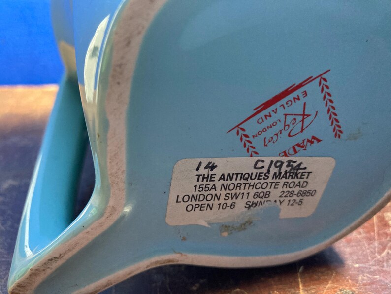 May include: A light blue ceramic pitcher with a white rim. The bottom of the pitcher has a sticker with text that reads "THE ANTIQUES MARKET" and "LONDON". The pitcher has a handle and a spout.