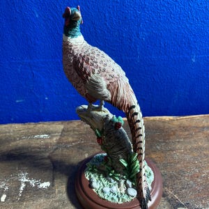 Può includere: Statuetta dettagliata di un fagiano appollaiato su un ramo. L'uccello ha piume marroni, bianche e nere, una testa rossa e un collo verde. La base è di forma circolare marrone con un tronco d'albero strutturato e fogliame verde.